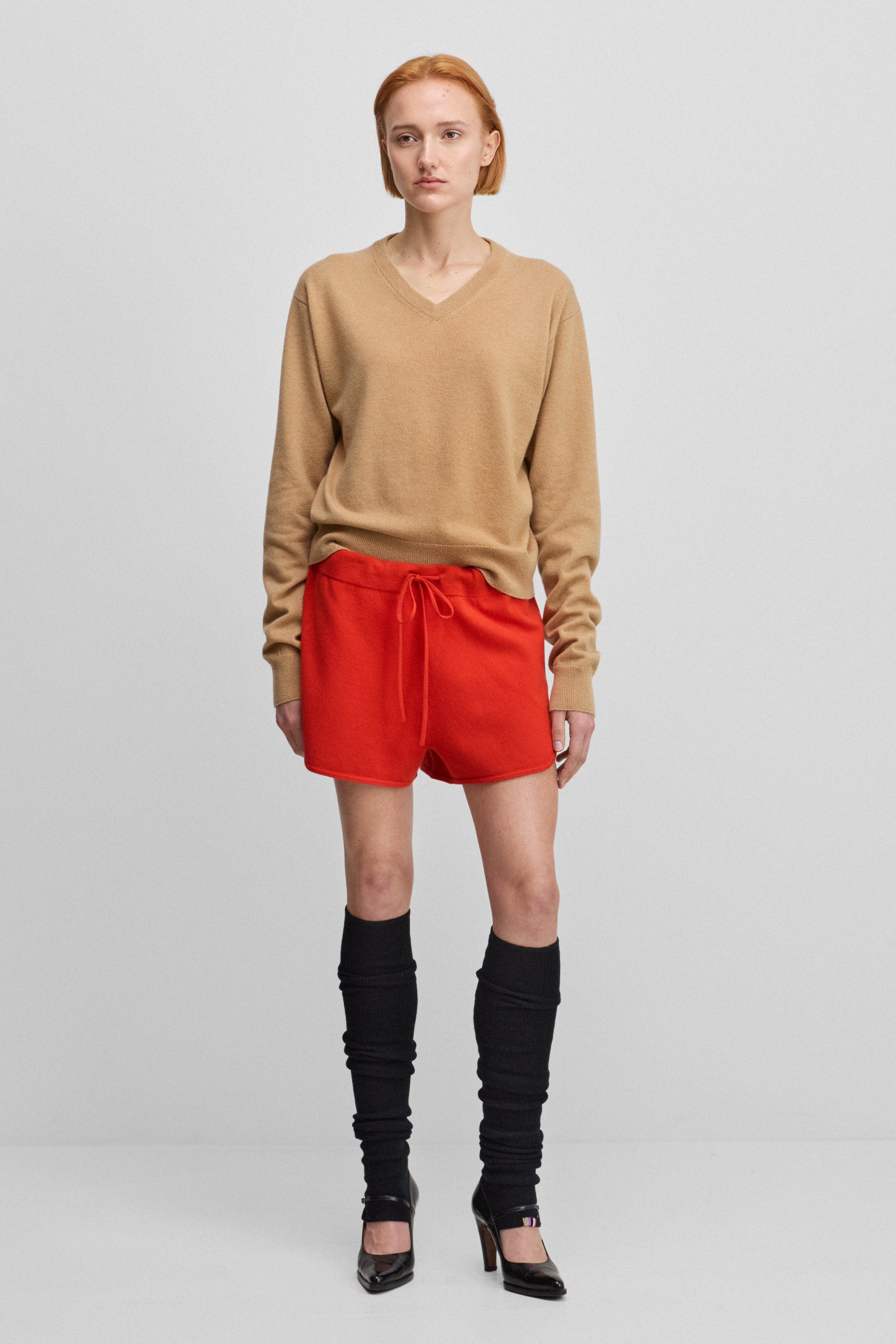 woman wearing a camel beige classic-fit cashmere v-neck sweater, styled with red shorts.
