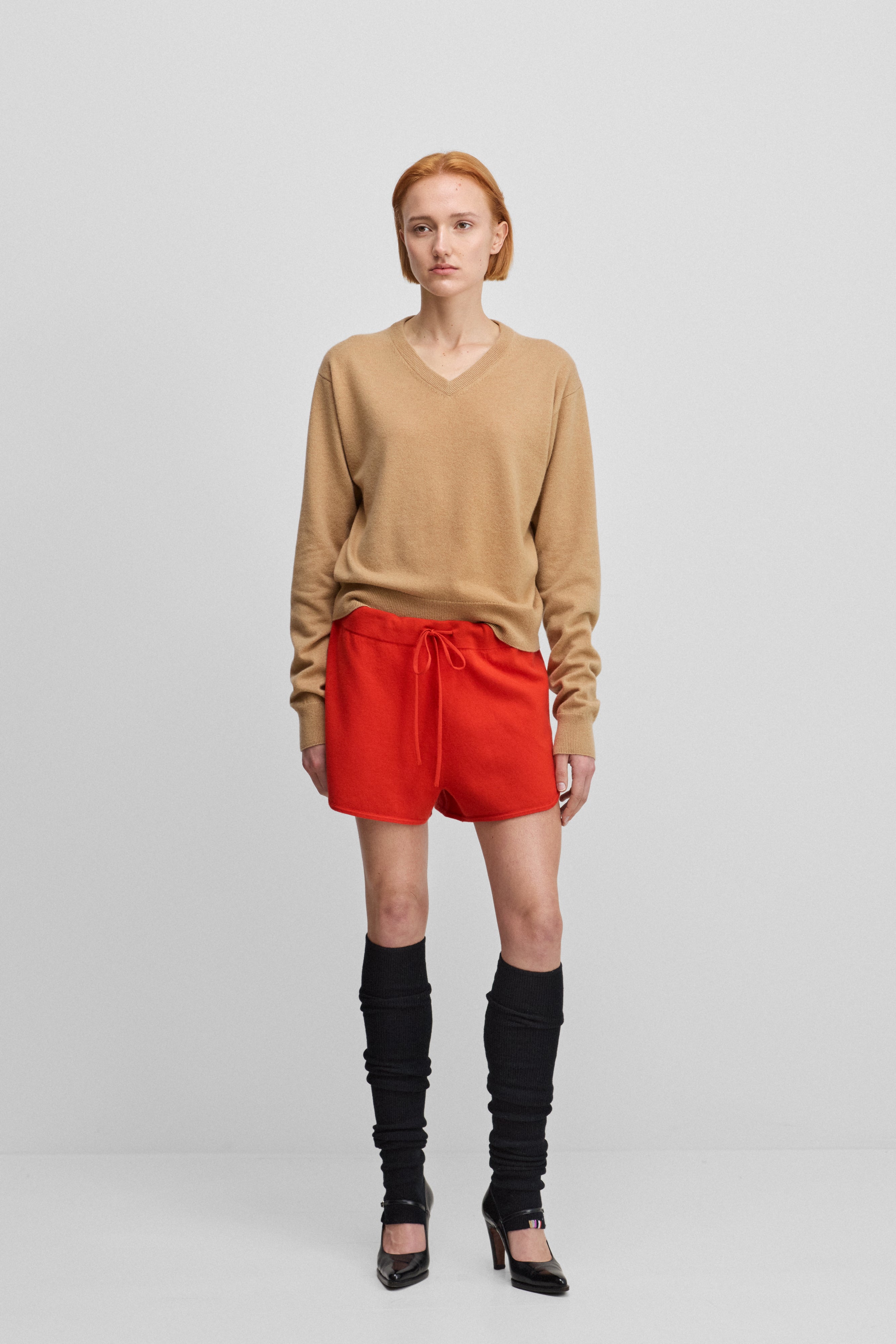 woman wearing a camel beige classic-fit cashmere v-neck sweater, styled with red shorts.