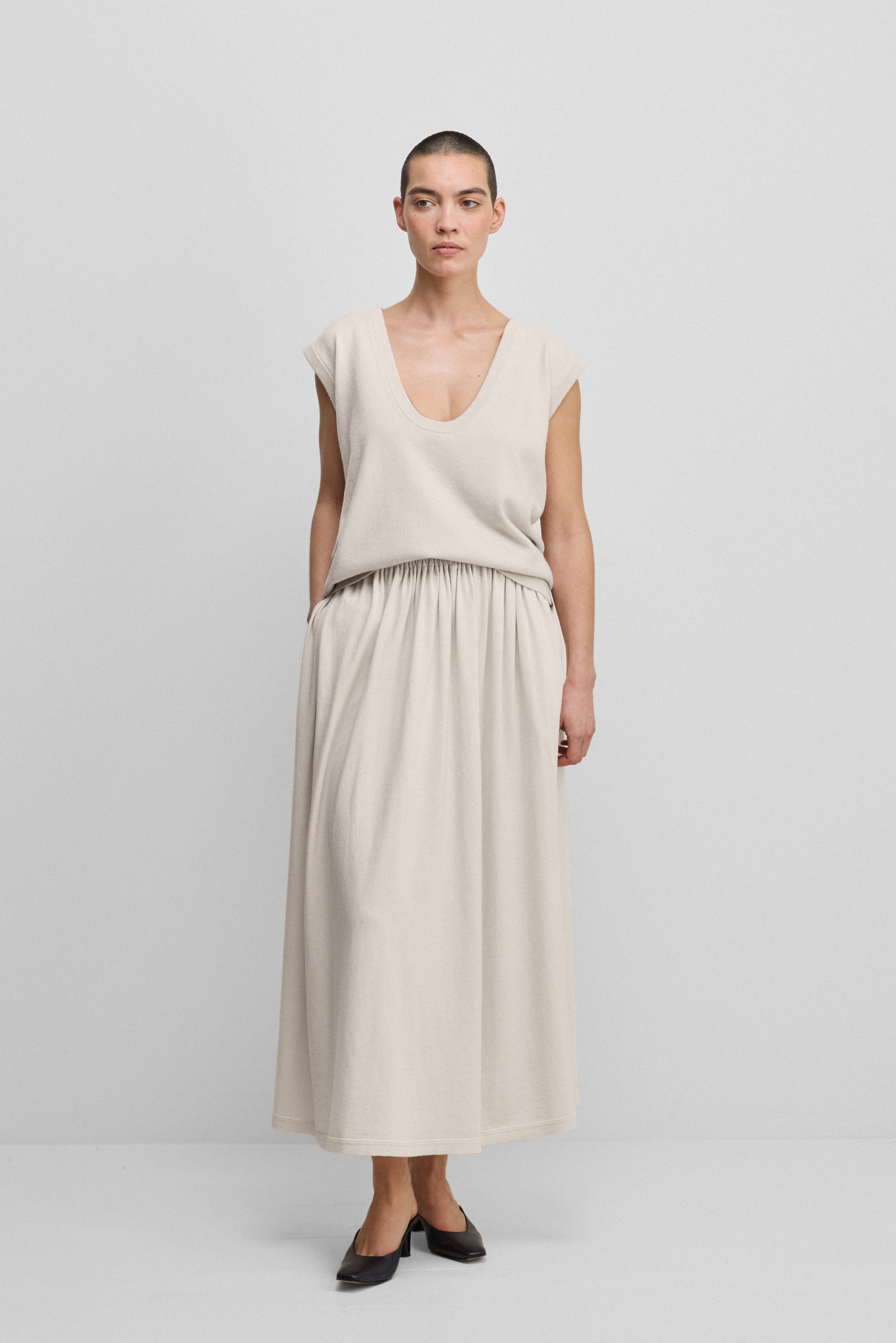 woman wearing an off-white sleeveless cashmere sweater vest with a scoop neck, styled with a white skirt.