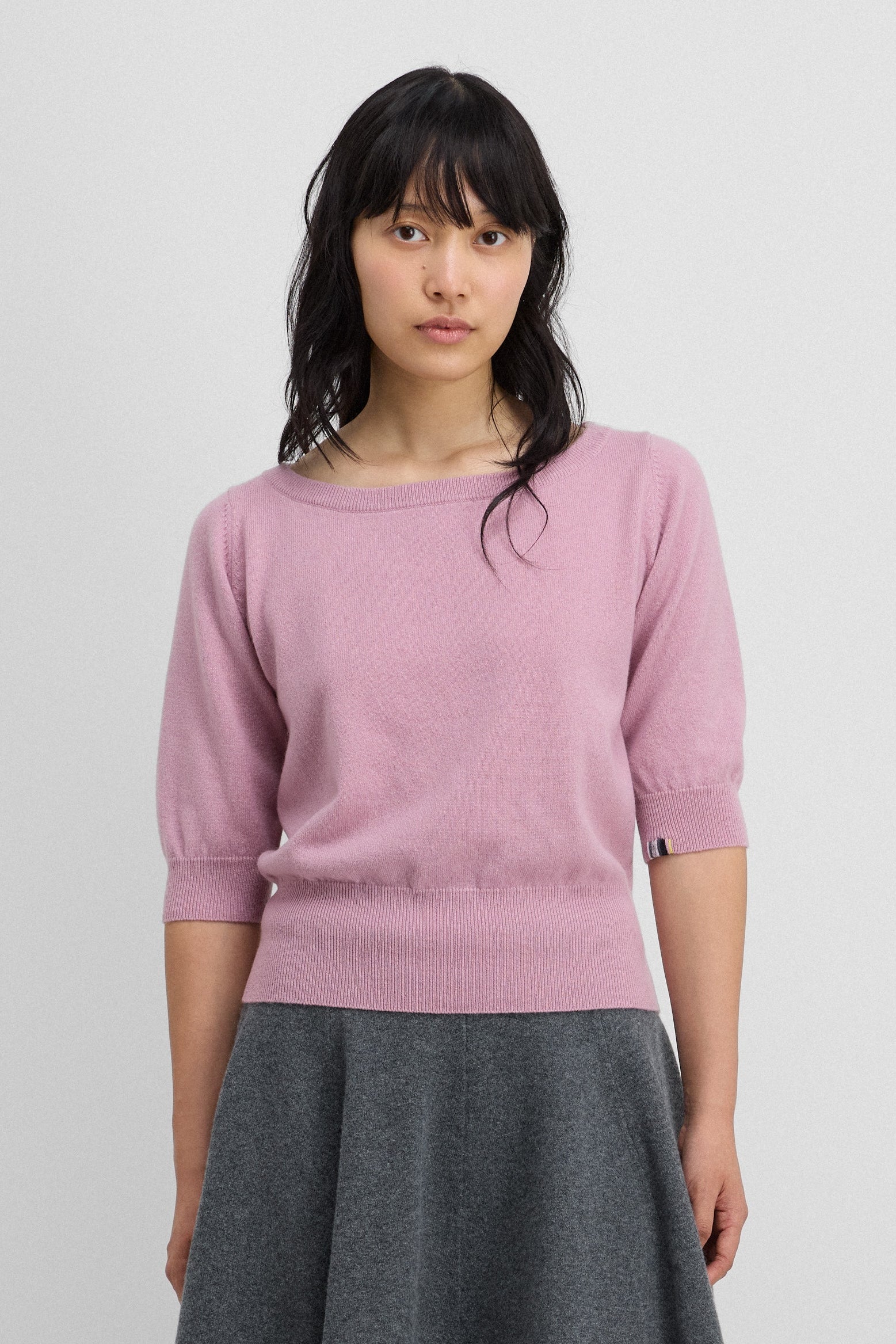 woman wearing a pink cashmere boat-neck sweater with elbow-length sleeves, styled with a skirt.